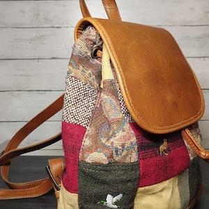May include: A backpack featuring a patchwork design of multi-colored woven fabric panels in shades of red, green, and tan, accented with a tan leather top flap, base, and straps. An embroidered white duck is visible on a dark green fabric panel.