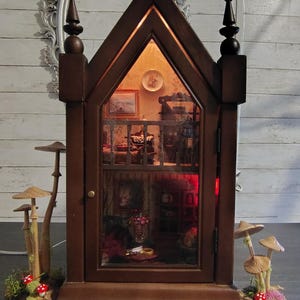 May include: A dark brown wooden gothic-style miniature cabinet with a pointed arch glass door, illuminated interior, and two levels of detailed room scenes. The cabinet is flanked by decorative clusters of tall wooden mushrooms and green moss.