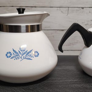 May include: Two white teapots of different sizes featuring a blue floral pattern on the front. Each teapot has a black handle, a black lid knob, and a silver-colored metal band around the upper section. The teapots have a rounded base shape.