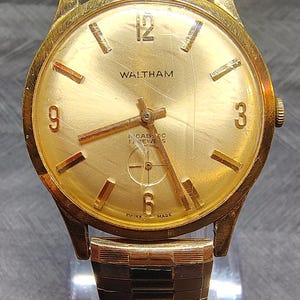 May include: A gold-tone Waltham wristwatch featuring a round gold dial with Arabic numerals at 3, 6, 9, and 12 o'clock, gold-tone hands, a sub-dial for seconds, and a textured gold-tone metal link bracelet. The dial is marked with Waltham and Swiss Made.