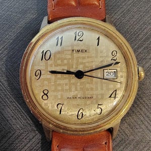 Könnte beinhalten: Eine Timex Armbanduhr mit goldenem, rundem Gehäuse und braunem Lederarmband. Das Zifferblatt hat ein strukturiertes Gittermuster, schwarze arabische Ziffern, schwarze Zeiger und ein Datumsfenster mit der Zahl 20. Der Text lautet Timex und Water Resistant.