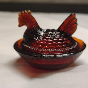 May include: A deep amber-coloured glass hen on a nest dish. The lid is shaped like a sitting hen with textured feather details, and the base is a round bowl with a cross-hatched pattern around the exterior rim, crafted from translucent glass.