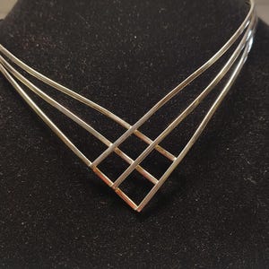 Może przedstawiać: Srebrzysty naszyjnik typu choker wykonany z metalu, składający się z trzech cienkich, warstwowych pasków, które zbiegają się w środku w geometryczny wzór kratki w kształcie litery V. Naszyjnik ma gładkie, polerowane wykończenie i sztywną konstrukcję.