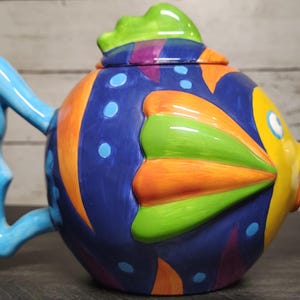 May include: A ceramic teapot shaped like a tropical fish with a vibrant blue, orange, yellow, and green color palette. The handle is light blue and shaped like a fin, while the spout features three white bubbles. A matching lid sits on top.