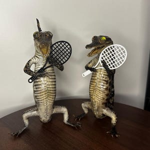 May include: Two taxidermy alligator figures standing upright on their hind legs. The left alligator holds a black plastic tennis racket, while the right alligator holds a white plastic tennis racket. Both creatures have open mouths and textured, scaly skin in shades of brown, tan, and cream.