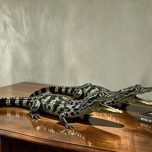 May include: Two realistic crocodile figurines with textured black and white scales, yellow eyes, and open mouths showing sharp teeth. The figures are positioned on a wooden surface next to a book titled Book of Taxidermy and a small analog clock.