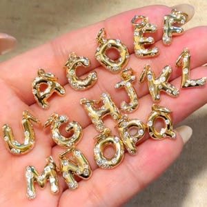 Op de afbeelding: Een verzameling letterbedels van goudkleurig metaal, elk met een afgerond, bol ontwerp ingelegd met kleine, heldere, sprankelende kristallen. Elke individuele letterbedel is voorzien van een klein metalen lusje aan de bovenkant.