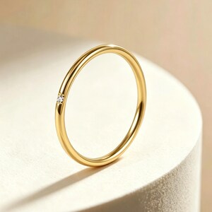 Op de afbeelding: Een dunne, gepolijste geelgouden ring met een enkele, kleine, ronde, heldere edelsteen die verzonken in het metaal is gezet. De ring heeft een glad, afgerond profiel en een hoogglans afwerking die het licht reflecteert langs de structuur.