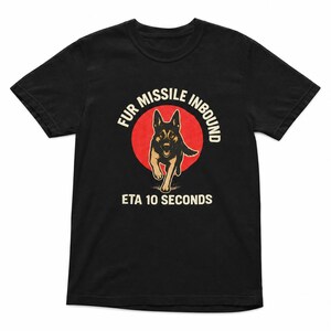 May include: A black short-sleeved t-shirt featuring a graphic of a running German Shepherd dog centered in front of a red circle. The text "FUR MISSILE INBOUND" is arched above the dog, and "ETA 10 SECONDS" is printed in white below the graphic.