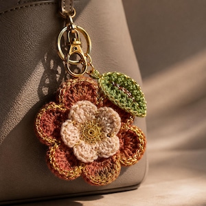 Op de afbeelding: Een gehaakte bloem-sleutelhanger met een gelaagd ontwerp met roestkleurige buitenste bloemblaadjes, lichtroze binnenste bloemblaadjes en een gouden hart. Een klein groen gehaakt blaadje is aan de zijkant bevestigd. De bloem zit aan een goudkleurige metalen karabijnhaak.