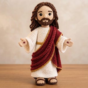 May include: A crochet doll depicting a figure with long brown hair and a beard, wearing a white robe with a dark red sash draped across the body. The figure has dark eyes, a tan complexion, and small red marks on the palms of the open hands.