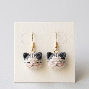 Op de afbeelding: Een paar oorbellen met kleine, gehaakte kattenkopjes van grijs en wit garen, met roze geborduurde wangen en gesloten ogen. De kattenkopjes zijn bevestigd aan goudkleurige metalen haken, wat zorgt voor een gedetailleerd en getextureerd accessoire.