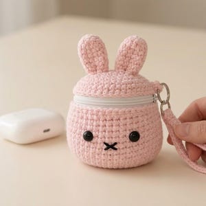 Può includere: Una custodia all'uncinetto rosa chiaro a forma di coniglio con due orecchie dritte, occhi a perla neri e una bocca a croce nera ricamata. La custodia presenta una chiusura a zip bianca, un anello in metallo argentato e un laccetto da polso.