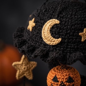 May include: A black crocheted dome-shaped ornament featuring a ruffled edge, decorated with embroidered tan crescent moons and stars. Dangling from the base are additional tan crocheted stars and a small orange crocheted jack-o'-lantern.