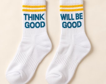 May include: A pair of white crew socks featuring two horizontal yellow stripes at the top. The left sock displays the dark teal text "THINK GOOD" and the right sock displays the dark teal text "WILL BE GOOD" in a bold, blocky font.