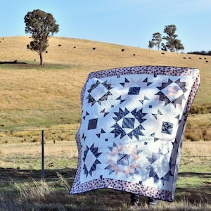 May include: A white quilt with a black and gray geometric pattern. The quilt is hanging on a fence in a field with a rolling hill in the background.