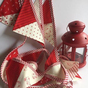 May include: A red and white fabric garland with a variety of patterns, including stripes, polka dots, and small designs. The garland is made of triangular flags and is hanging in a loop. A red lantern is in the background.