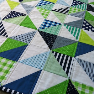 Op de afbeelding: Een patchworkdeken met een geometrisch patroon van driehoeken in tinten groen, blauw en wit. De deken is gemaakt van verschillende stoffen met verschillende patronen, waaronder stippen, strepen en ruitjes.