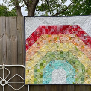 May include: A colorful handmade quilt with a rainbow design. The quilt features a spectrum of colors, including red, orange, yellow, green, and blue, arranged in an arched pattern. The quilt is hanging on a wooden fence.
