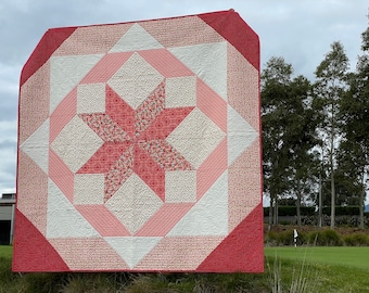 May include: A red, pink, and white quilt with a star pattern in the center. The quilt is hanging on a wooden post in front of a green grassy field.