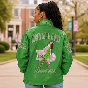May include: A green windbreaker jacket featuring the text Alpha Kappa Alpha, 1908, and Pretty Girl. The back displays a graphic of a person in a green dress and pink hat, surrounded by white pearls and the number 08, with AKA and SP 29 on sleeves.