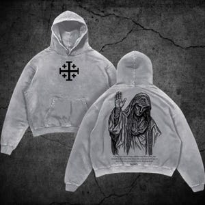May include: A grey hooded sweatshirt featuring a black Jerusalem cross on the front chest. The back displays a detailed black graphic illustration of a robed figure with a raised hand, accompanied by printed text below the image.