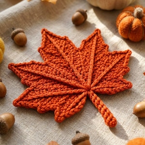 May include: A burnt orange crochet maple leaf with a textured, raised vein pattern. The leaf is shaped with five distinct points and a central stem, showcasing a dense, hand-stitched yarn construction that creates a tactile, dimensional surface.