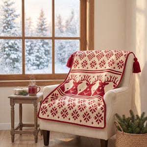May include: A cream-colored knit throw blanket featuring a repeating red pattern of reindeer and snowflakes, finished with red tassels at the top corners. The blanket is draped over a cream-colored armchair positioned near a window.