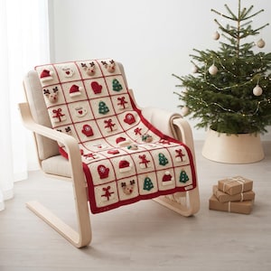 May include: A cream-colored crochet throw blanket featuring a grid of square patches with raised, three-dimensional holiday motifs including reindeer, Christmas trees, gift boxes, mittens, Santa hats, and mugs, all bordered by a red yarn trim.