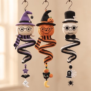 May include: Three hanging crochet ornaments featuring a witch, a pumpkin, and a skeleton. Each figure has a spiral body, round glasses, and a hat. The witch has a purple hat, the pumpkin has a black hat, and the skeleton wears a black top hat.