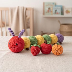 Può includere: Un bruco giocattolo all'uncinetto con testa rossa, antenne a spirale viola e corpo segmentato giallo, verde e viola. Accanto ci sono tre piccoli oggetti all'uncinetto: una fragola rossa, una mela rossa e un pallone da basket arancione.