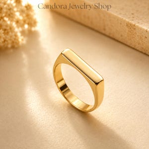 Op de afbeelding: Een goudkleurige ring met een glad, rechthoekig bovenoppervlak. De ring is gepolijst en reflecterend, vangt het licht op en benadrukt de strakke, solide constructie. De tekst Candora Jewelry Shop is zichtbaar in het bovenste gedeelte.