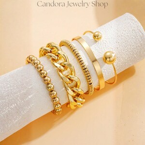 May include: A set of four gold-toned rings displayed on a white cylindrical holder. The collection includes a beaded ring, a thick chunky chain link ring, a textured rope-style ring, and an open-ended cuff ring featuring two polished gold spheres.
