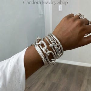 Könnte beinhalten: Ein Stapel silberfarbener Armbänder am Handgelenk, bestehend aus einer breiten, gehämmerten Manschette, einer groben ovalen Gliederkette, einem Perlenarmband und einem glatten, polierten Armreif. Das Set enthält einen passenden Silberring.