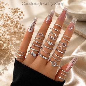 May include: A collection of silver-toned rings worn on fingers, featuring various designs including hearts, wavy bands, twisted textures, and small embedded clear stones. The text Candora Jewelry Shop is visible at the top of the image.