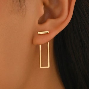May include: A gold-toned ear jacket earring featuring a small horizontal bar stud that sits on the front of the earlobe and a larger, open rectangular frame that hangs behind the earlobe, creating a layered and geometric appearance.