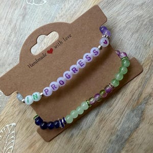 May include: Two beaded bracelets displayed on a brown cardboard tag. The top bracelet features white beads with purple letters spelling IN PROGRESS and a small heart icon. The bottom bracelet consists of green, purple, and dark blue beads.