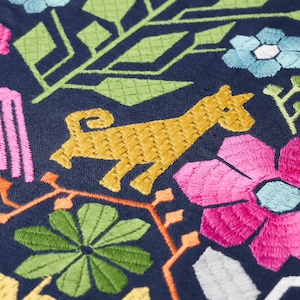 Pode incluir: Close-up de tecido bordado com fundo azul marinho. O design apresenta um animal amarelo, flores rosa e azuis, folhas verdes e vinhas laranja. O bordado &eacute; detalhado e colorido, criando um t&ecirc;xtil vibrante.
