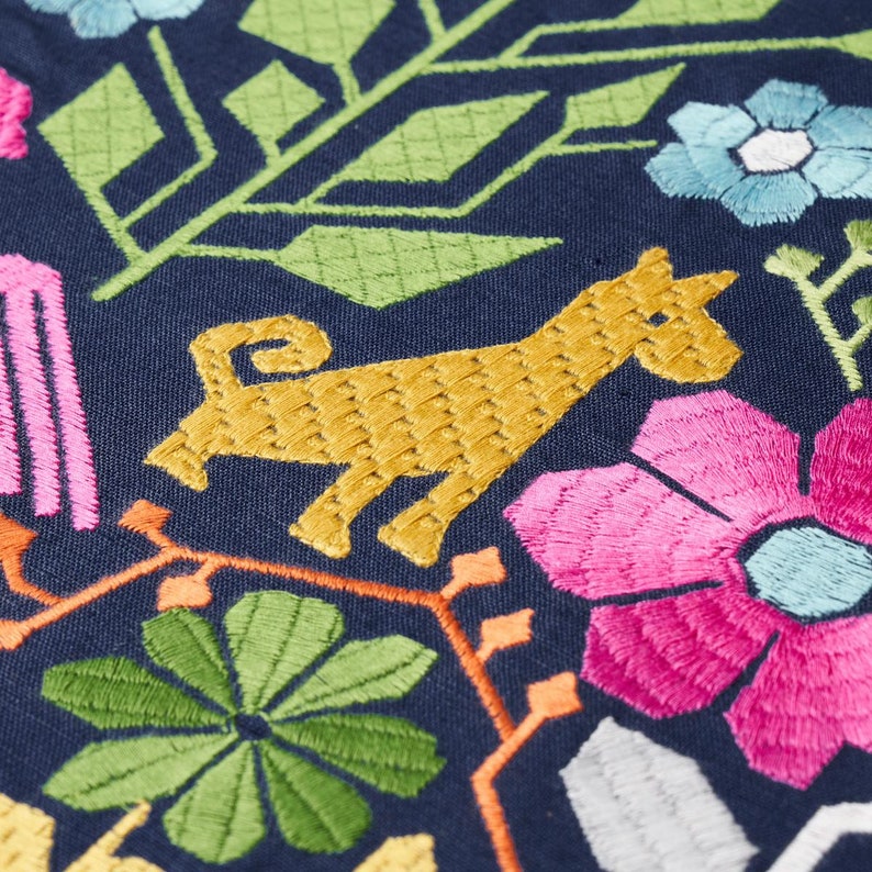 Pode incluir: Close-up de tecido bordado com fundo azul marinho. O design apresenta um animal amarelo, flores rosa e azuis, folhas verdes e vinhas laranja. O bordado &eacute; detalhado e colorido, criando um t&ecirc;xtil vibrante.