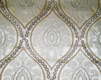 May include: A close-up of a cream-colored fabric with a repeating pattern of embroidered silver and brown swirls and circles.