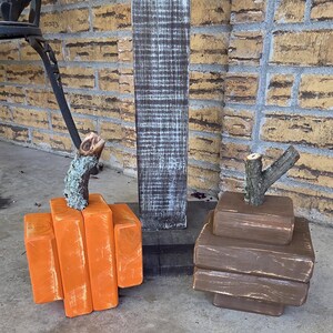 May include: Two decorative pumpkins crafted from stacked wooden blocks. One pumpkin is painted bright orange with a distressed finish, and the other is painted brown. Both feature natural, textured tree branch stems attached to the top.