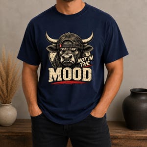 May include: A navy blue t-shirt featuring a graphic of a bull wearing a baseball cap. The bull has a nose ring and a stern expression. Below the bull, the text reads "NOT IN THE MOOD" in a distressed, blocky font with a red horizontal line accent.