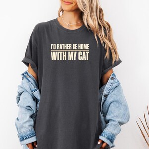 Peut inclure: Un t-shirt gris anthracite à manches courtes avec le texte crème "I'D RATHER BE HOME WITH MY CAT" imprimé sur la poitrine. Le t-shirt est porté sous une veste en jean bleu clair aux manches retroussées, créant un style superposé décontracté.