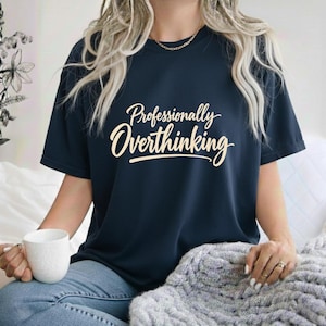 May include: A dark navy blue short-sleeve t-shirt featuring the text "Professionally Overthinking" printed in a light cream-colored script font across the chest. The shirt has a crew neckline and is worn with blue denim jeans.