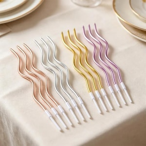Könnte beinhalten: Ein Set aus zwölf langen, dünnen, gewellten Geburtstagskerzen in vier Farbgruppen: Roségold, Silber, Gold und Lila. Jede Kerze hat eine metallische Oberfläche und ist auf einem kleinen, weißen Kunststoffsockel befestigt.
