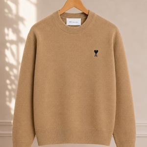 May include: A tan long-sleeve crewneck sweater featuring a ribbed collar, cuffs, and hem. The chest displays a small, black embroidered heart-shaped logo with the letter A underneath. A white interior neck label reads ami alexandre mattiussi.