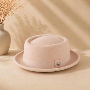 Op de afbeelding: Een lichtbeige vilten pork pie hoed met een bijpassende grosgrain lintband. De hoed heeft een platte kroon met een cirkelvormige inkeping en een klein zeshoekig metalen embleem aan de zijkant van de band, voorzien van een subtiel bijenmotief.