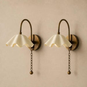 Peut inclure: Une paire d'appliques murales dotées de bras incurvés en laiton antique et de supports muraux circulaires. Chaque lampe possède un abat-jour en forme de cloche, couleur crème et ondulé, ainsi qu'une chaînette métallique avec une petite bille.