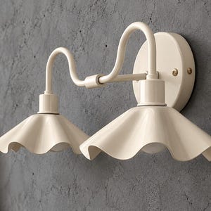 Op de afbeelding: Een crèmekleurige wandlamp met twee gebogen armen die uit een ronde basis komen. Elke arm ondersteunt een brede, gegolfde, klokvormige kap die het licht naar beneden richt, wat zorgt voor een symmetrisch en functioneel verlichtingsontwerp.