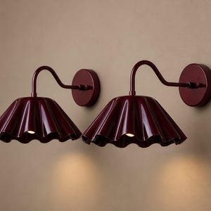 Peut inclure: Une paire d'appliques murales bordeaux foncé dotées d'un abat-jour brillant en forme de cloche festonnée, fixé à un bras incurvé et à une plaque de montage circulaire. Chaque luminaire diffuse une lumière chaude sous le bord métallique cannelé.